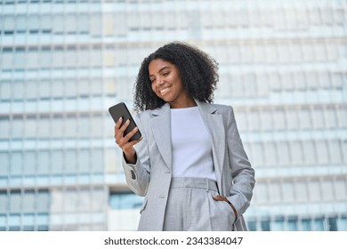 Happy young African American business woman using cell phone outdoor. Smiling lady professional holding smartphone standing at city street building looking at mobile working outside office. - Powered by Shutterstock