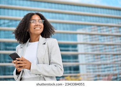 Happy young African American business woman using cell mobile phone outdoors. Professional lady holding cellphone standing at city street looking away working on smartphone outside office, copy space.