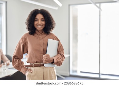 Happy young African American business woman standing in office. Smiling confident professional female company employee leader manager holding laptop looking at camera. Portrait. Copy space. - Powered by Shutterstock