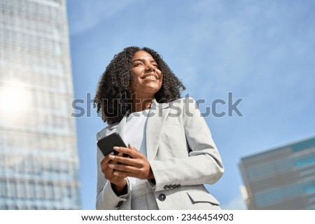 Happy young African American business woman holding smartphone walking in city street, smiling ethnic lady using financial app on cell phone looking away with mobile tech working outside office.