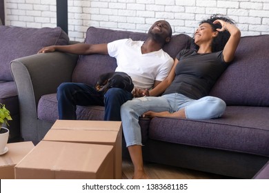 Happy Young African American Couple Rest On Cozy Couch Together With Favorite Pet Companion, Have Break Relocating To New Home, Excited Black Family With Dog Relax On Sofa Glad To Move In Own House