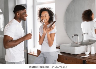 Happy Young African American Couple Brushing Teeth, Wife Showing Her Smartphone To Husband, Having Fun While Engaging In Dental Care Routine In Modern Bathroom Indoors, Free Space - Powered by Shutterstock