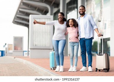 Happy Young African American Family Standing With Suitcases Near Airport Terminal Or Railway Station, Trying To Catch Taxi After Arrival To New Destination, Lady Raising Hand With Thumb Up To Stop Cab