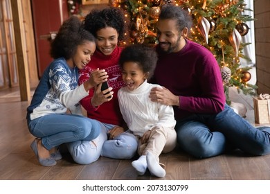 Happy Young African American Family With Two Small Kids Relax Near Christmas Tree Have Fun With Modern Cellphone. Smiling Biracial Parents With Little Children Laugh Use Smartphone Gadget Together.