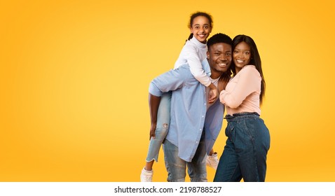 Happy Young African American Family Posing, Man Giving Piggy Back Ride For His Daughter, Looking And Smiling Together At Camera Over Yellow Background, Panorama With Free Space