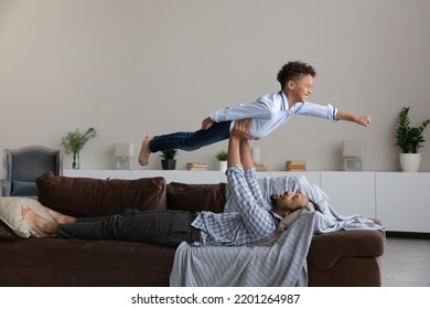 Happy Young African American Father Lifting In Air Laughing Adorable Little Child Son, Lying On Comfortable Sofa At Home. Happy Two Male Generations Family Having Fun, Enjoying Entertaining Activity.