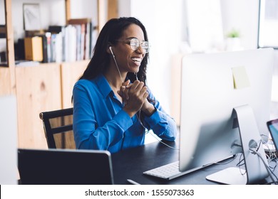 Happy Young African American Female Office Worker In Blue Shirt And Glasses Sitting At Work Table With  Hands Together Wearing Headphone And Conducting Online Negotiations With Coworkers