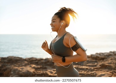 Happy young african american female in sports wear, headphones with smartphone running in morning on rocks near ocean alone, outdoor. Health care, training, active lifestyle and sports with music - Powered by Shutterstock
