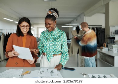 Happy young African American female fashion designer pointing at paper with sketch held by colleague during discussion of new items - Powered by Shutterstock