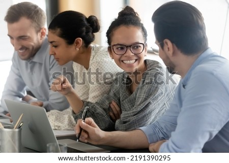 Happy young african american female employee in eyeglasses enjoying discussing project ideas with male colleague, working in small group on computers in office, teamwork collaboration concept.