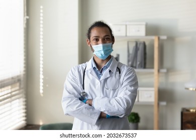 Happy Young African American General Practitioner Doctor Therapist In White Medical Uniform Wearing Protective Respirator Facemask Standing With Folded Arms In Clinic Office, Healthcare Concept.