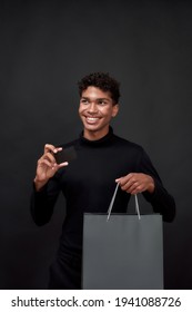 Happy Young African American Guy With Paper Bag Holding Credit Card Mockup And Looking To Side While Posing On Dark Background, Vertical Shot. Discount And Sale Concept