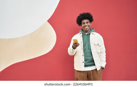 Happy young African American guy standing at red wall outdoors using cell phone, looking at camera holding cellphone enjoying doing online ecommerce shopping in mobile apps, chatting or playing games. - Powered by Shutterstock