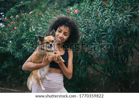Happy young African American lady with curly hair in sport outfit hugging adorable little chihuahua against green plants in garden