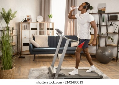 Happy young african american male athlete in headphones drinking bottled water enjoying favorite song while running on treadmill during intensive workout at modern apartment. - Powered by Shutterstock