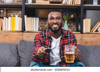 Happy Young African American Man Holding Glass Of Beer And Remote Controller While Sitting On Sofa At Home