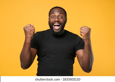 Happy Young African American Man 20s In Casual Basic Black T-shirt Isolated On Yellow Background Studio Portrait. People Sincere Emotions Lifestyle Concept. Mock Up Copy Space. Doing Winner Gesture