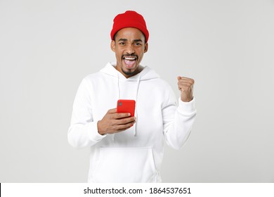 Happy Young African American Man 20s In Basic Casual Streetwear Hoodie Standing Using Mobile Cell Phone Typing Sms Message Doing Winner Gesture Isolated On White Colour Background, Studio Portrait