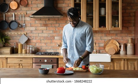 Happy Young African American Man Chopping Fresh Vegetables On Wooden Board In Kitchen, Preparing Vegetarian Health Meal. Smiling 30s Multiracial Mixed Race Guy Involved In Cooking Alone At Home.