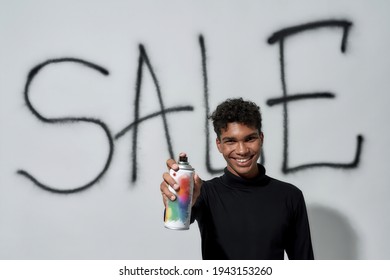 Happy Young African American Man Holding Paint Sprayer While Standing On White Wall With Sale Inscription On Background And Looking At Camera. Discount And Sale Concept