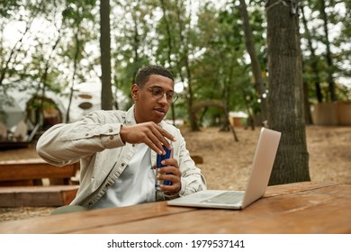 Happy Young African American Man Opening Can With Energy Drink While Looking Into Laptop Screen Outdoors. Work In Nature Concept