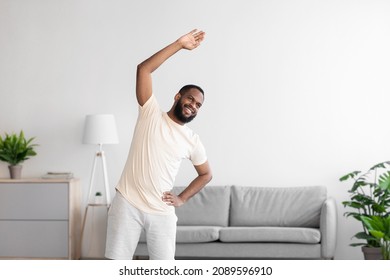 Happy young african american man with beard in white sportswear doing exercise and warm-up in living room interior. Sports, body care, weight loss and workout during covid-19 pandemic, copy space - Shutterstock ID 2089596910