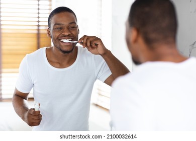 Happy Young African American Man Brushing His Teeth With Toothbrush In Bathroom, Handsome African American Guy Smiling At Mirror, Enjoying Making Morning Dental Hygiene, Selective Focus On Reflection