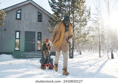 Happy young African American man pulling sledge with cute boy while his wife pushing their son on back during winter stroll in the country - Powered by Shutterstock