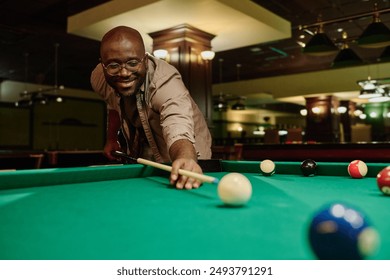 Happy young African American man with billiard cue looking at ball and hitting it while bending over pool table and playing at leisure