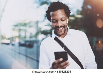 Happy young African American man in headphone walking at sunny city and making video conversation with friends on his electronic smart phone.Blurred background