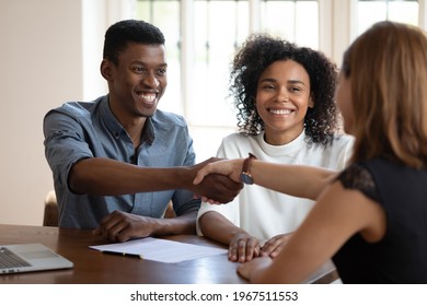Happy young african american married couple clients shaking hands with financial advisor, discussing contract terms of conditions. Emotional glad millennial diverse spouses celebrating making deal.