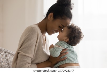 Happy Young African American Mom Hug Cuddle With Little Baby Kid Daughter At Home. Smiling Mixed Race Ethnicity Mother Embrace Small Ethnic Newborn Toddler Child Show Love Care. Motherhood Concept.