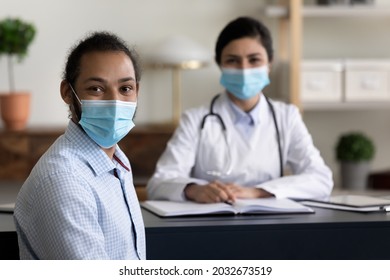 Happy Young African American Patient In Protective Facemask Looking At Camera, Posing In Clinic Office With Confident Indian Ethnicity Female Doctor In Respirator On Background, Healthcare Concept.