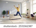 Happy young African American woman in yellow jumper and jeans spends weekend at home, feels full of energy and jumps up in living room with sofa, rug, clothes rack, plants, shelves and desk with