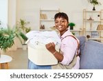 Happy young African American woman sitting on couch with basket full of washed clothes, looking cheerful at camera. Smiling black housewife rejoicing with clean laundry in living room at home.