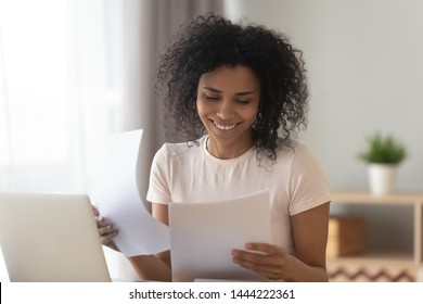 Happy Young African American Woman Sit At Table Reading Good News In Paper Letter Checking Domestic Bills, Smiling Black Holding Documents Doing Paperwork Work Or Study Sit At Home Office Desk