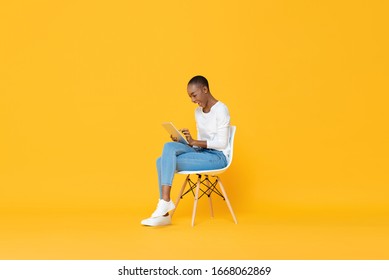 Happy Young African American Woman Sitting On A Chair Using Tablet Computer In Yellow Isolated Studio Background