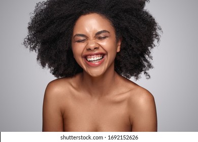 Happy young African American woman with bare shoulders laughing against gray background while representing cosmetology and skincare industry