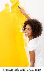 Happy Young African American Woman With A Cute Curly Afro Hairstyle Painting A Wall Orange With A Roller As She Does Home Renovations