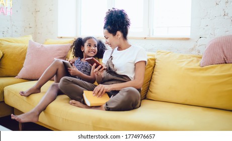 Happy Young African American Woman With Smartphone And Notebook Laughing With Playful Little Daughter With Tablet While Sitting On Yellow Sofa In Light Modern Living Room