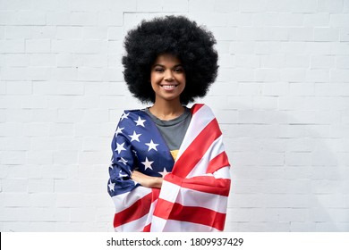 Happy young African American woman with Afro hair standing on white background wrapped in USA flag looking at camera outdoor. Independent confident smiling mixed race lady headshot portrait.