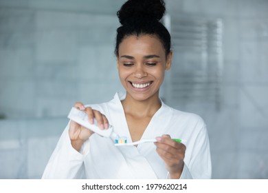 Happy young African American woman squeezing mint toothpaste from tube on plastic toothbrush for brushing teeth in bathroom, keeping morning routine for dental care, healthy enamel, mouth hygiene