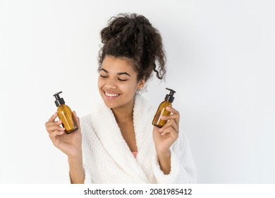 Happy young african american woman in bathrobe holding glass bottles with cosmetics, compare products, choose essential oil for anti-aging therapy, smiling wide, stand on white copy space background