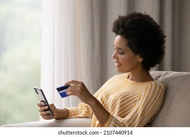 Happy young African American woman holding cellphone and bank credit card in hands, transferring money online, shopping goods in internet store, purchasing services, satisfied with secure payments. - Shutterstock ID 2107440914