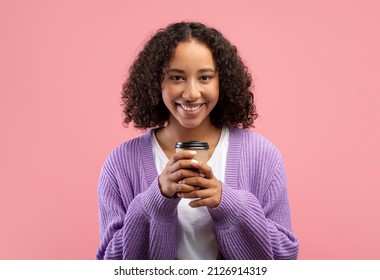 Happy young African American woman holding takeaway coffee in disposable paper cup and smiling at camera on pink studio background. Lovely black lady making break with hot takeout drink