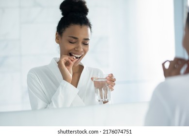 Happy young African American woman taking pill of vitamin, food supplement, drinking fresh water, holding glass in bathroom. Healthcare, pharmacy, wellness concept. Mirror reflection - Powered by Shutterstock