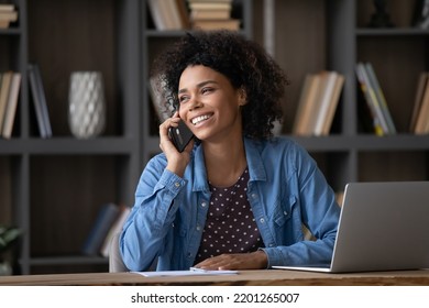 Happy Young African American Woman Involved In Mobile Phone Call Conversation, Distracted From Computer Work, Discussing Issues Distantly, Listening Good News, Sitting At Table At Home Office.