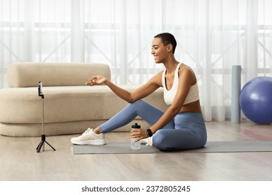 Happy young african american woman blogger in sportswear sit on mat, shooting video fitness blog on phone in living room interior. Health care, weight loss and sports at home, fit lifestyle - Powered by Shutterstock