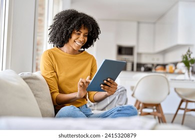 Happy young African American woman sitting on couch at home using digital tablet. Smiling relaxed lady relaxing on sofa looking at tab technology device holding pad computer in hands in living room. - Powered by Shutterstock