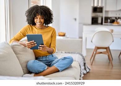 Happy young African American woman sitting on couch at home using digital tablet. Smiling relaxed lady relaxing on sofa looking at tab technology device holding pad computer in hands. Copy space - Powered by Shutterstock